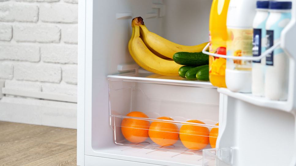Close up of a mini fridge, filled with fresh bananas, zucchini, oranges, juice and milk