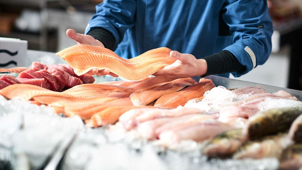 A close up of hands holding a long slice of fresh fish. An assortment of fresh, raw fish are set out on ice at a fish market.